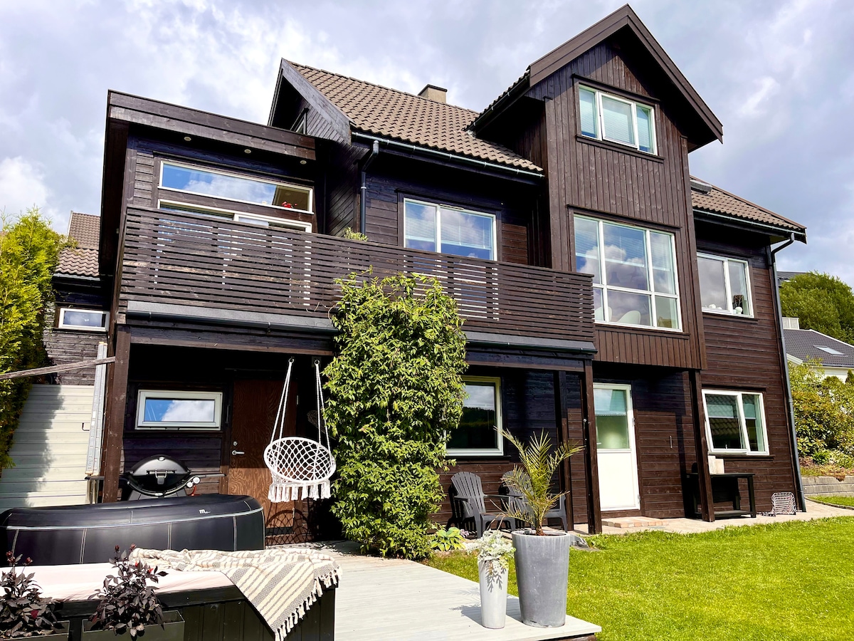 A charming two-story wooden house features a large balcony adorned with greenery. A comfortable hanging chair and potted plants can be seen on the outdoor deck, alongside a barbecue grill. Large windows allow for ample natural light and provide views of the surrounding landscape.