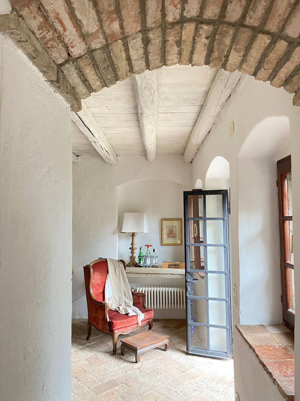 A cozy nook is displayed with a vintage red armchair positioned near a small desk. The room features a rustic ceiling with exposed wooden beams and terracotta flooring. Natural light filters through an open glass door, enhancing the warm and inviting ambiance.