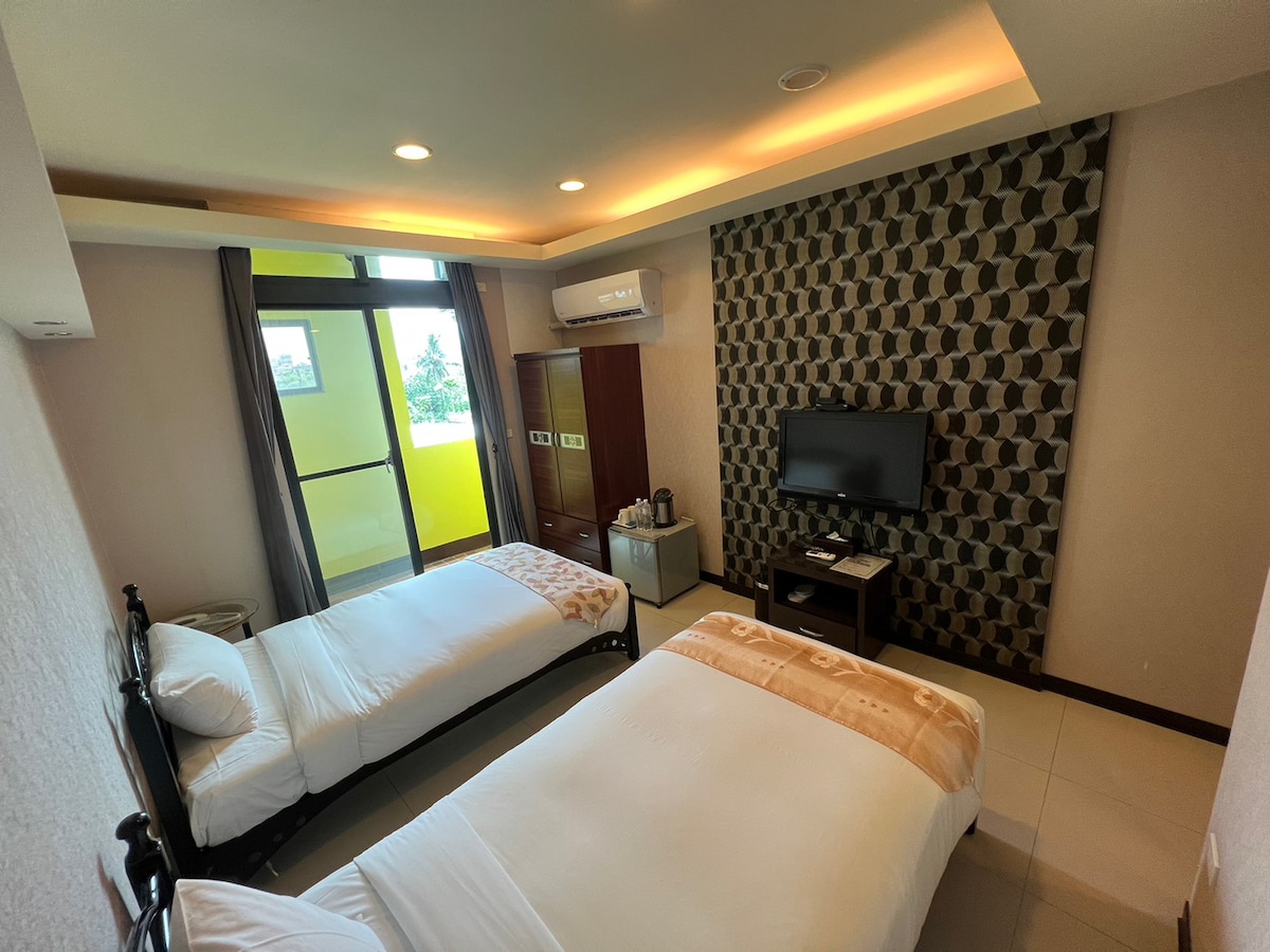 A spacious double room features two single beds dressed in white linens, set against a patterned accent wall. Natural light enters through large windows, illuminating the room. A flat-screen television and basic amenities are visible, providing comfort and convenience.