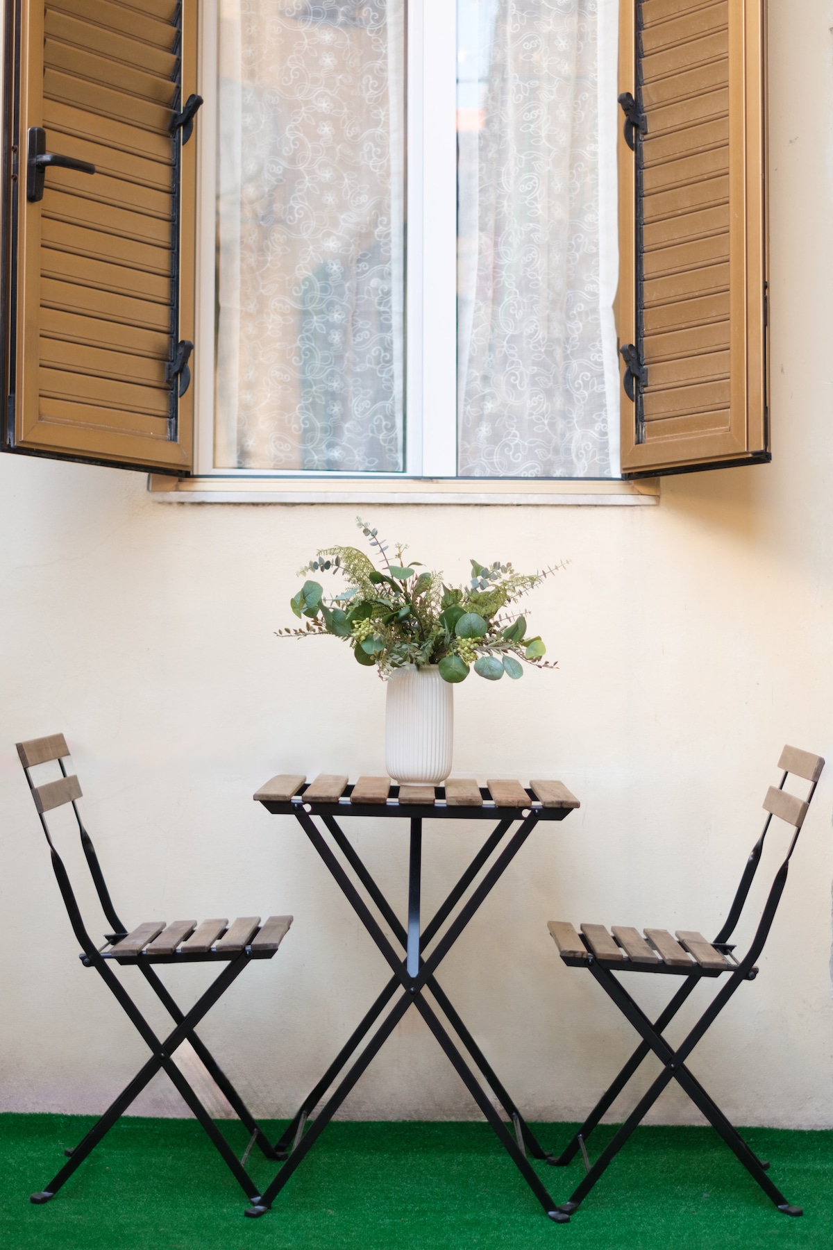An outdoor space features a small table flanked by two folding chairs, all set against a backdrop of a beige wall. A vase with greenery sits on the table, while a window with open wooden shutters allows natural light to illuminate the area.