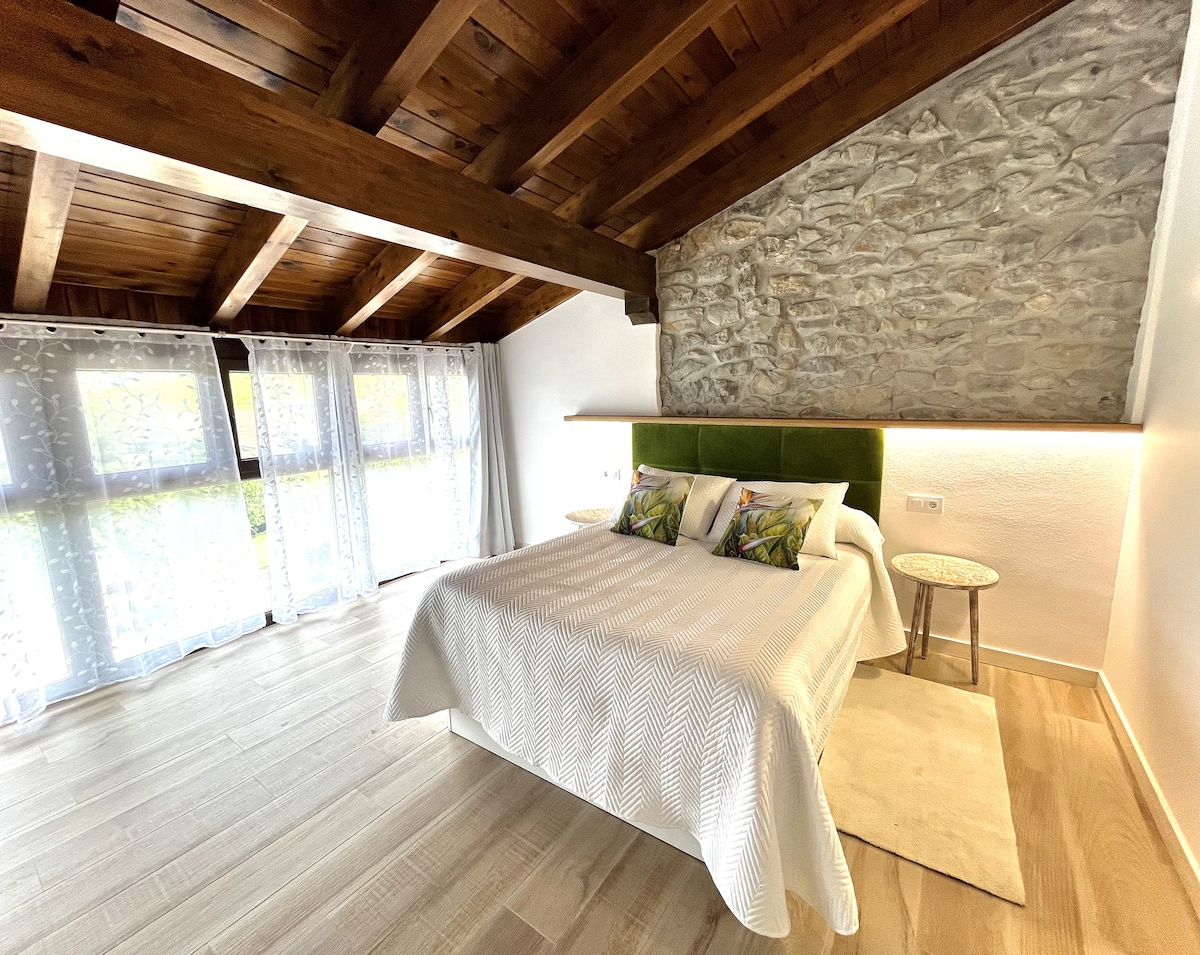 A spacious bedroom features a textured stone wall complemented by polished wooden beams. A large bed with a light-colored quilted cover is flanked by side tables. Natural light streams through expansive windows adorned with sheer curtains, illuminating the serene space.