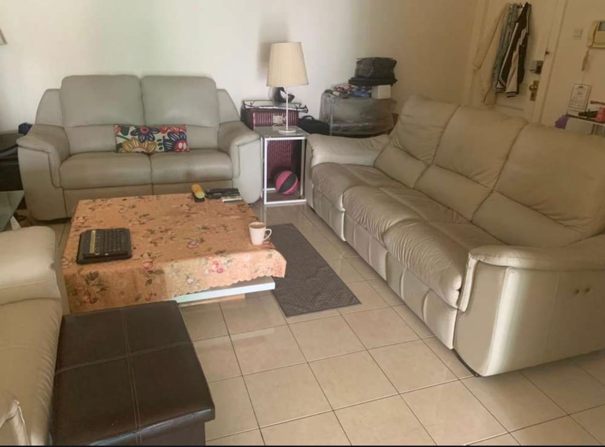 A comfortable living area is presented with two light-colored sofas facing each other. A floral-patterned coffee table sits in the center, accompanied by a small dark ottoman. A lamp on a side table provides lighting, while a neutral tile floor enhances the space.