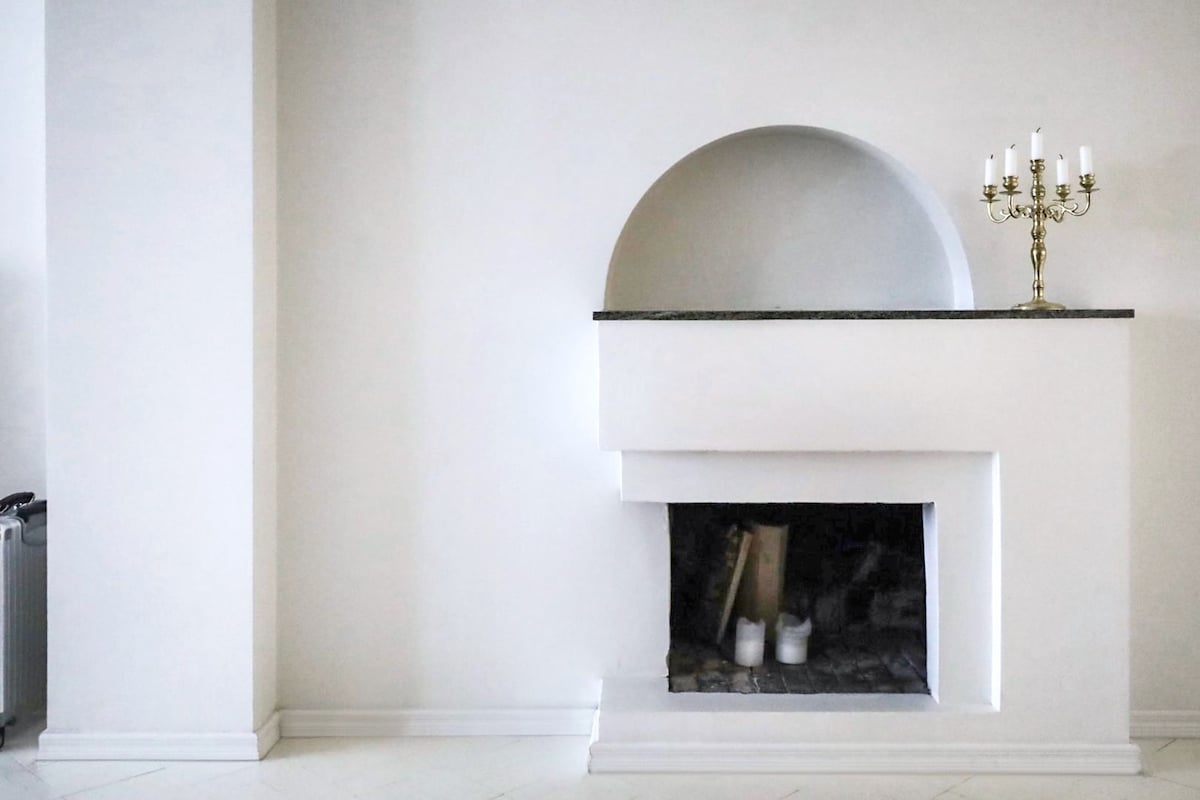 A minimalist fireplace is set within a white wall, featuring a curved alcove above a black granite ledge. A candelabra with white candles is positioned on the ledge, and logs are visible inside the fireplace. The surrounding space is clean and bright.