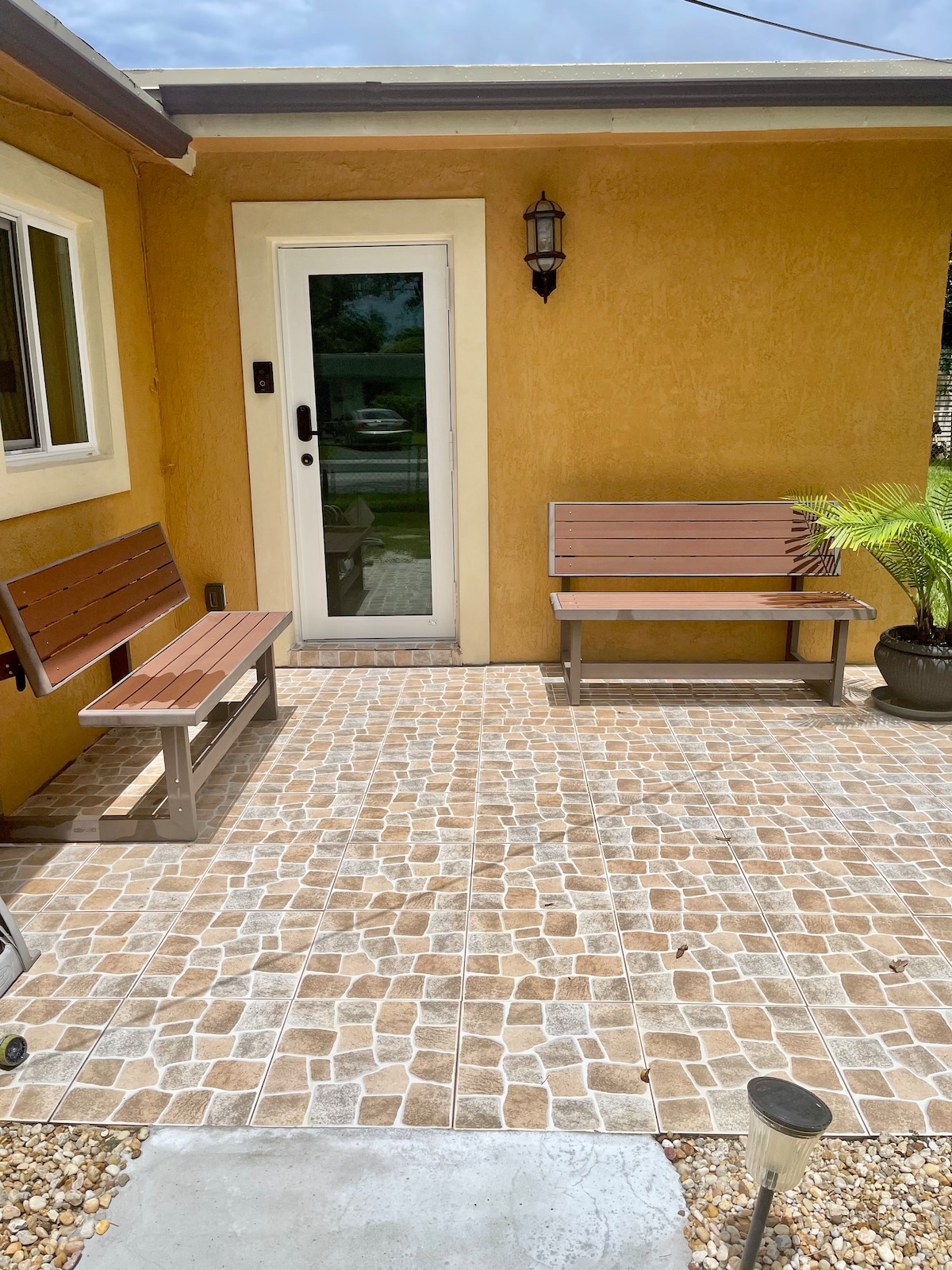 A small outdoor patio area is visible, featuring two wooden benches and a potted plant. The surface is composed of patterned tiles, complemented by a door leading to the guesthouse. Soft lighting is provided by a lantern mounted on the wall.