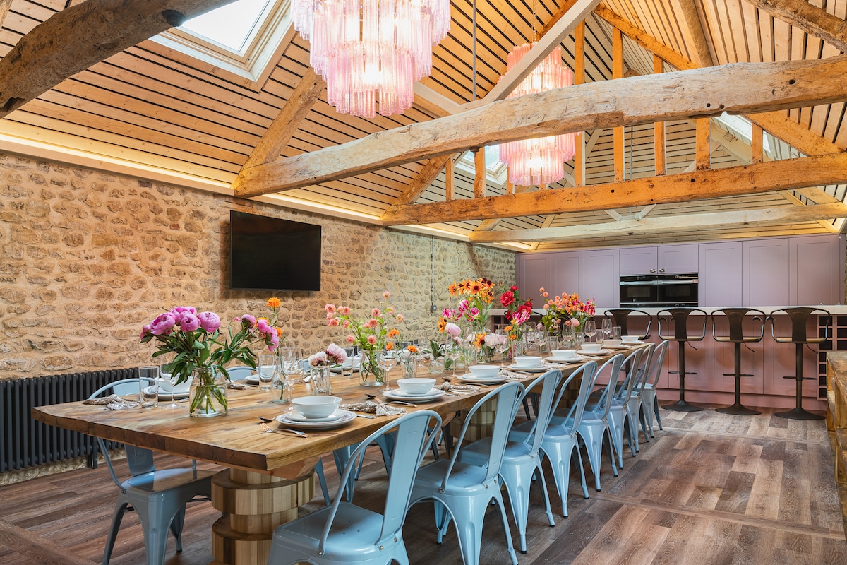 A spacious dining area features a long wooden table surrounded by light blue chairs. Colorful floral arrangements adorn the table, complemented by elegant tableware. Exposed wooden beams span the ceiling, with pink pendant lights adding a modern touch. A kitchen area with subtle cabinetry is visible in the background.