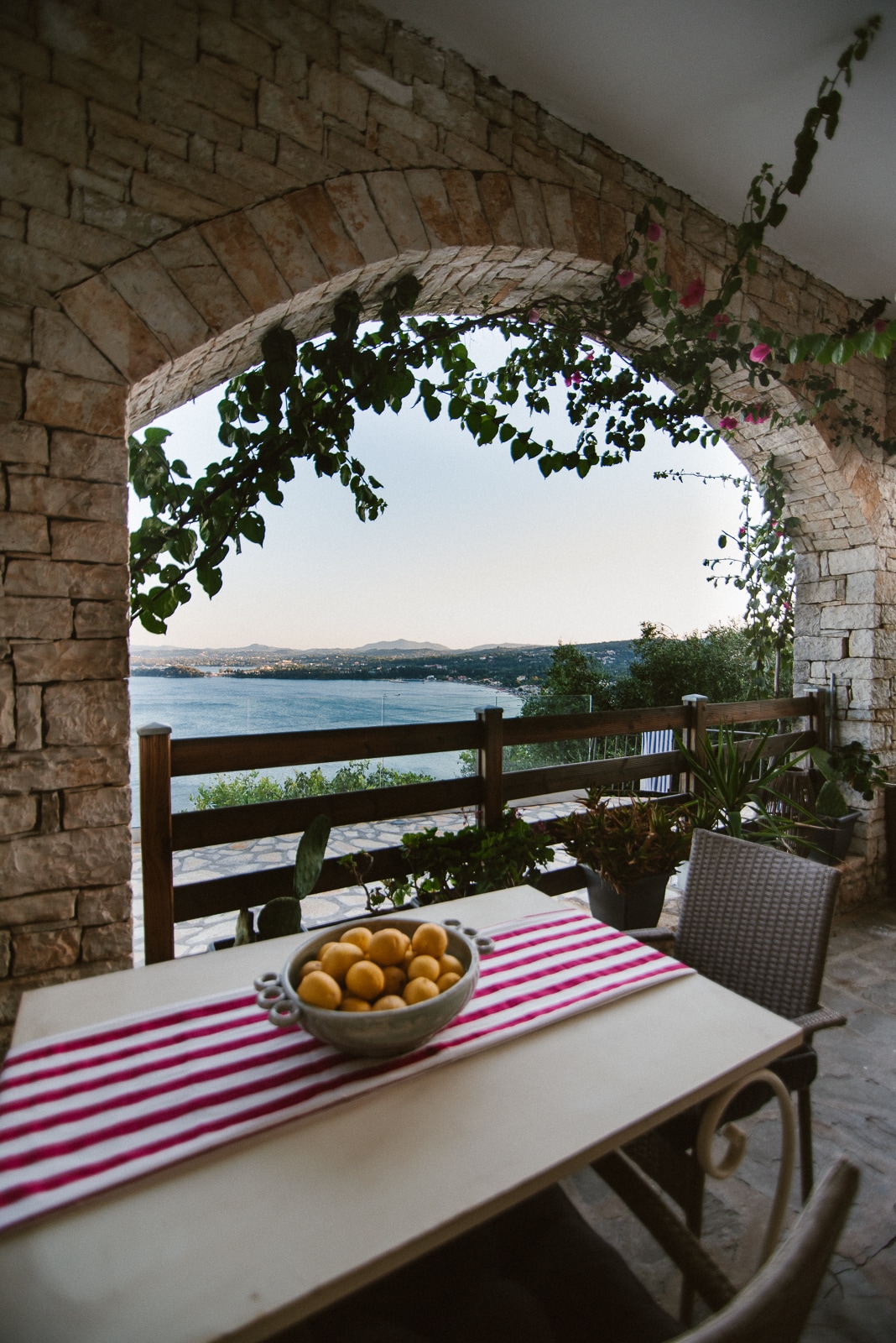 An arched stone doorway frames a serene outdoor terrace, showcasing a wooden table adorned with a bowl of lemons. A patterned table runner contrasts with the natural stone, while lush greenery cascades from above, offering a glimpse of the tranquil sea and distant hills.