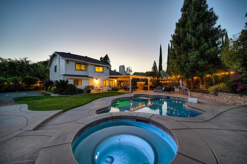 An inviting backyard features a spacious swimming pool and hot tub, both illuminated with soft lighting. Lush greenery surrounds the area, complemented by a well-maintained lawn. The home stands in the background, with exterior lights enhancing the serene evening atmosphere.