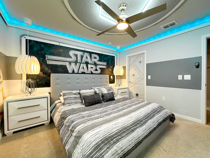 Elegantné útočisko v štýle Star Wars s manželskou posteľou s geometrickou posteľnou bielizňou, obklopenou zodpovedajúcimi nočnými stolíkmi a sochárskymi lampami. Figúrka BB-8 a zarámované umelecké dielo zvýrazňujú sivé zatemňovacie závesy. Výnimočné vlastnosti LED osvetlenia