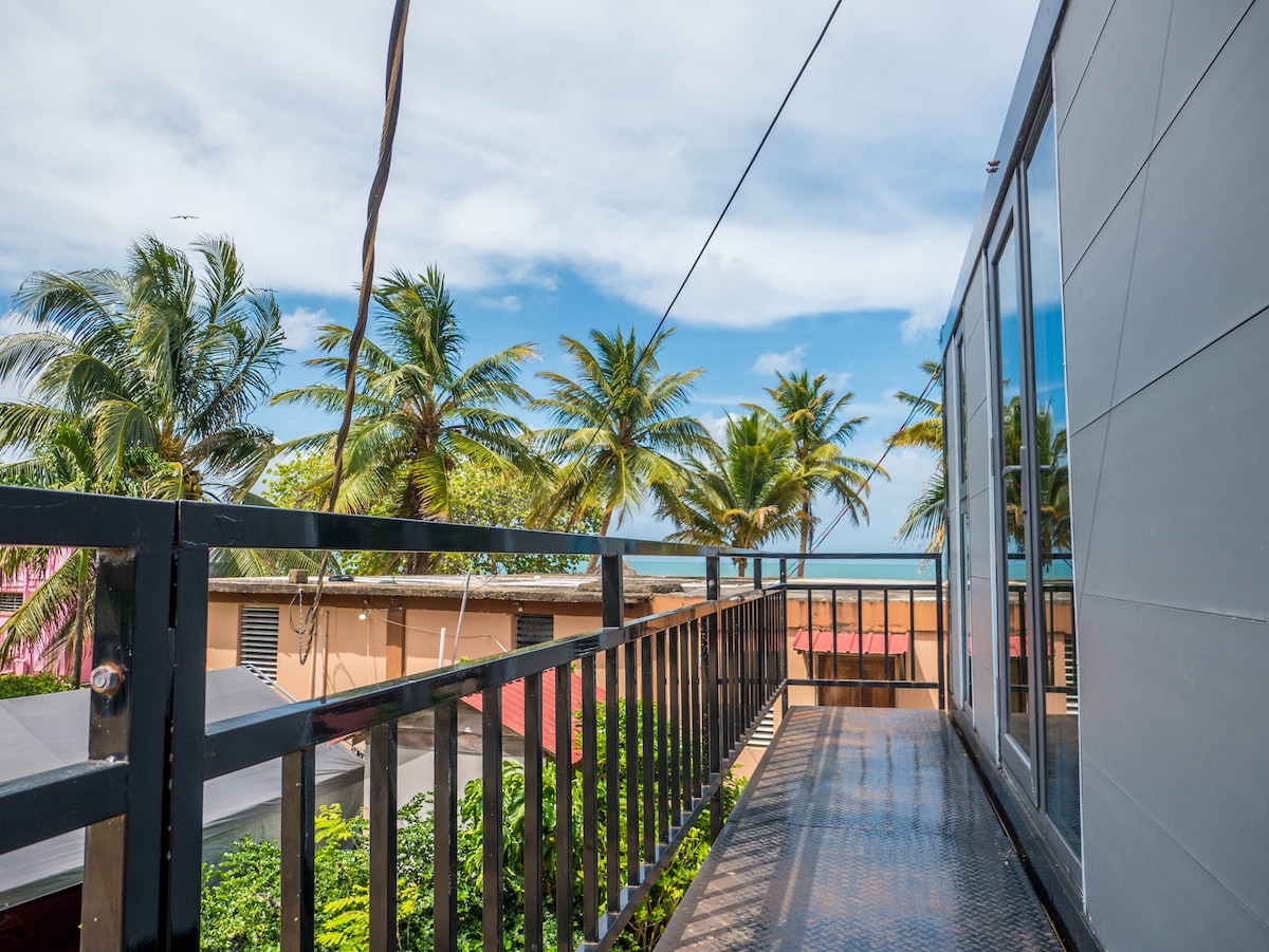 A metal walkway leads to an expansive balcony framed by palm trees, providing a view of the Caribbean Sea and surrounding rooftops. Clear skies above contribute to a serene atmosphere, enhancing the outdoor experience.