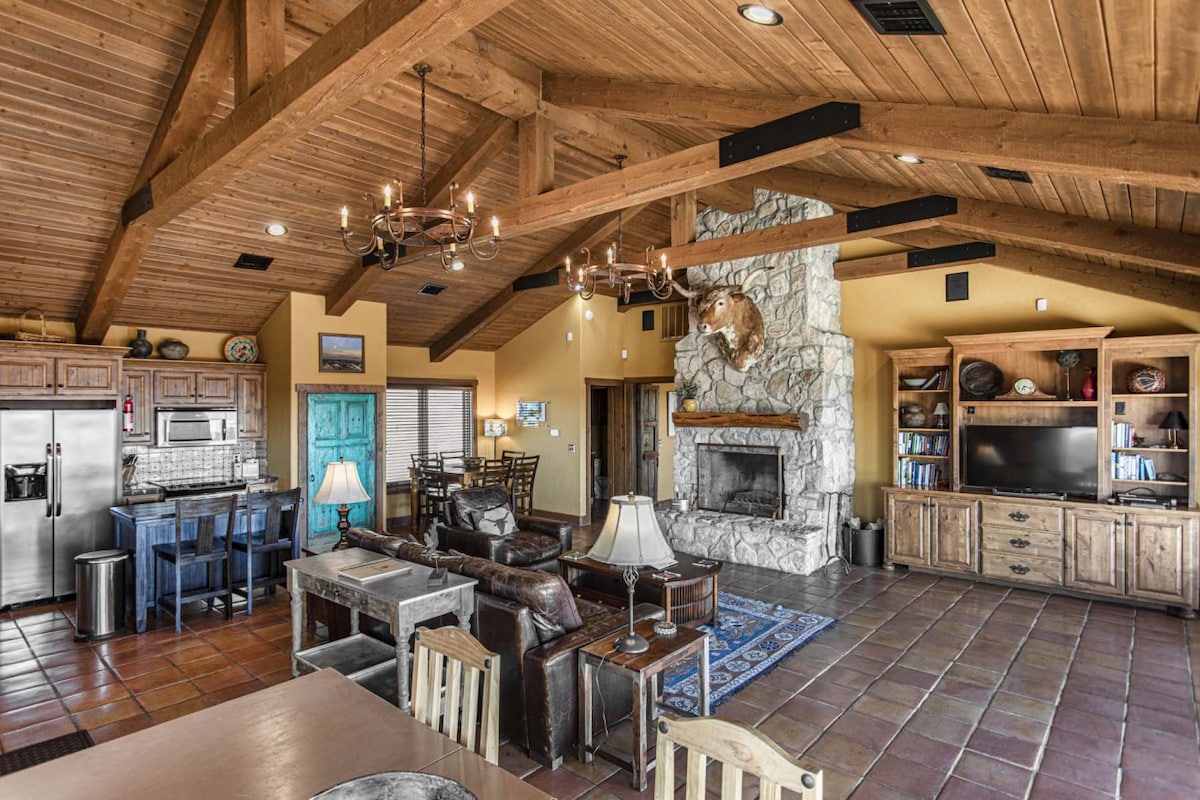 An open living area features a stone fireplace, exposed wooden beams, and ample natural light. Leather seating is arranged around a central coffee table, complemented by a decorative rug. A dining area and kitchen with modern appliances are visible in the background.