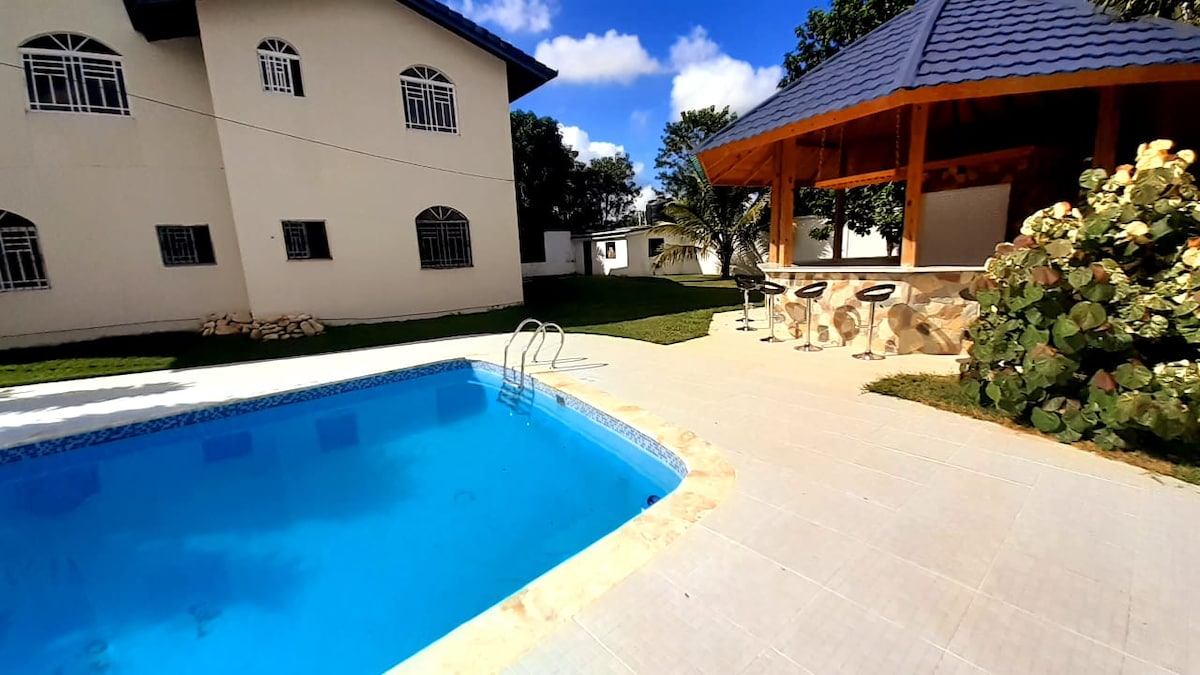 A sparkling pool is surrounded by a tiled patio, complemented by a kiosk featuring bar seating. Lush greenery adds a natural touch, while a spacious building stands in the background. Clear blue skies create a bright and inviting atmosphere.