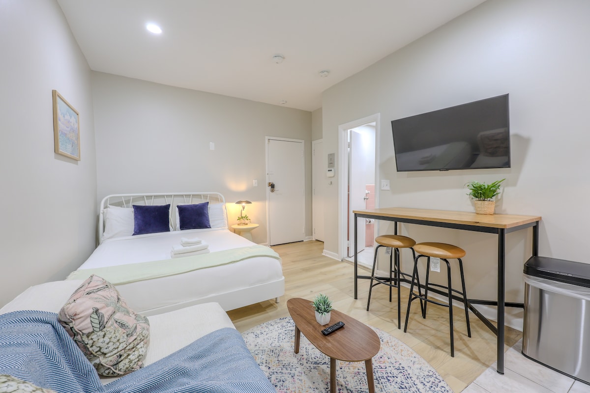 A studio apartment is shown with a spacious bed dressed in white linens, complemented by decorative pillows. A compact dining table with three stools is positioned near a wall-mounted television. A cozy seating area features a light-colored sofa and a small coffee table, creating an inviting atmosphere.