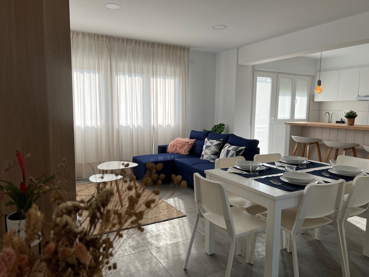 A modern living area is showcased, featuring a blue sectional sofa adorned with decorative pillows. A dining table with white chairs is set for a meal, while natural light filters through sheer curtains covering multiple windows.