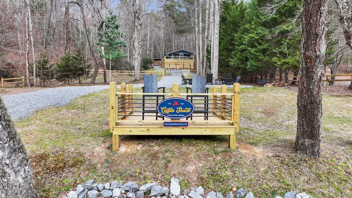 A wooden deck is featured at the forefront, offering two POLYWOOD Adirondack chairs and a welcoming sign for the Caddis Shack. The surrounding area is characterized by natural vegetation and gravel paths, leading to the cabin visible in the background, set among tall trees.