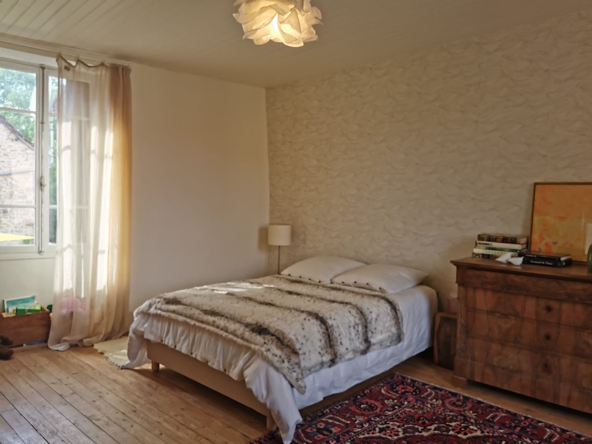 A spacious bedroom is presented with a large bed covered in a soft blanket. Natural light fills the room through a window with sheer curtains. A wooden dresser and a small lamp are positioned nearby, adding a calming touch to the decor.