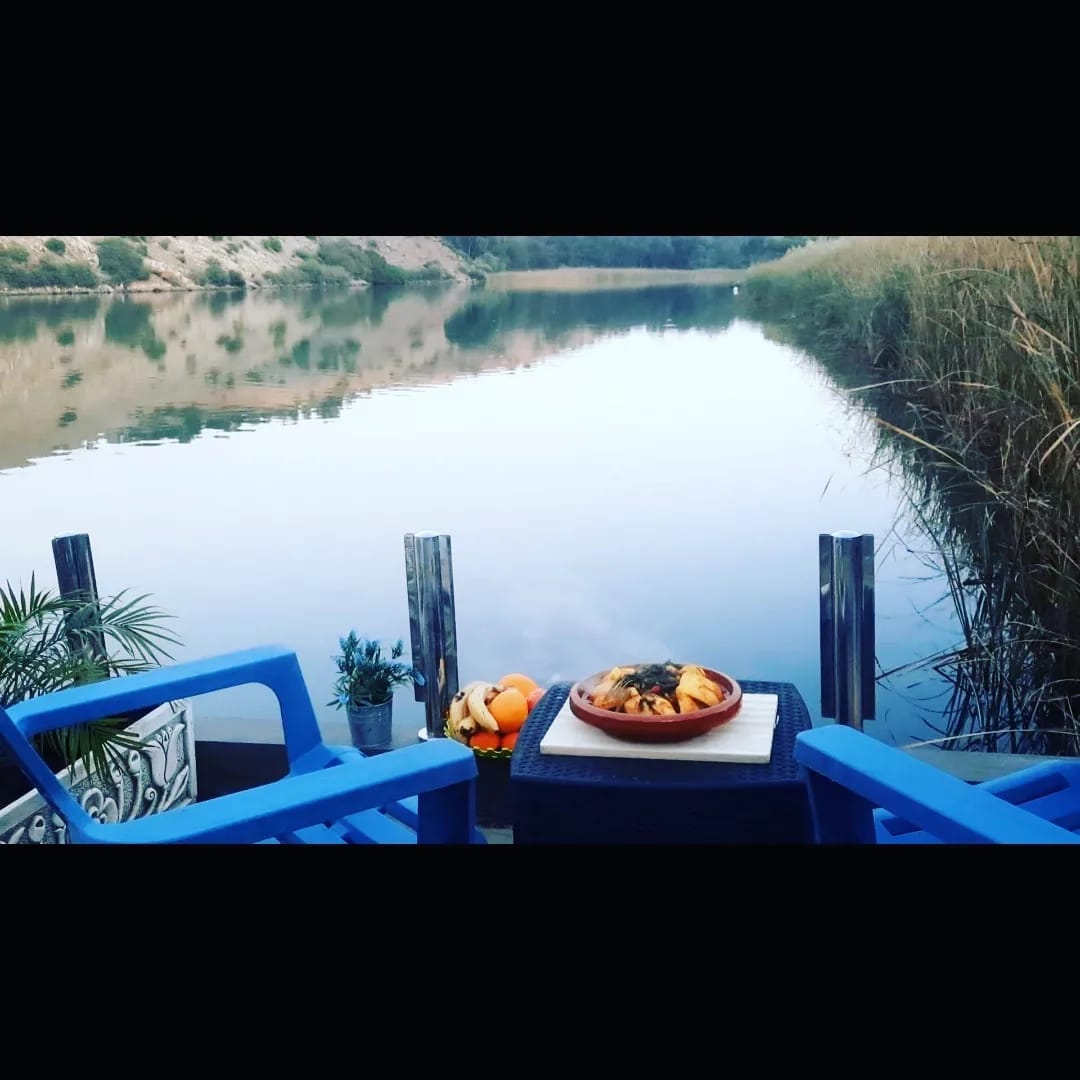 A peaceful riverside scene showcases a table set with a colorful dish, accompanied by fresh fruit. Two blue chairs offer comfortable seating, while lush greenery frames the view of calm waters reflecting the surrounding nature.