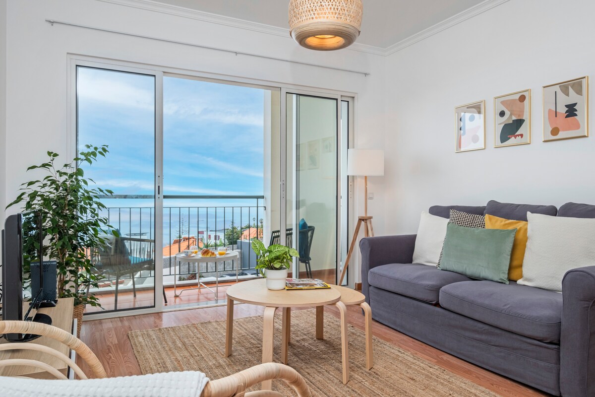 A bright living area features a comfortable sofa with an assortment of cushions. A small round table rests in front, complemented by a plant. Large glass doors provide access to a balcony with ocean views, while light filters in from the window, enhancing the serene ambiance.