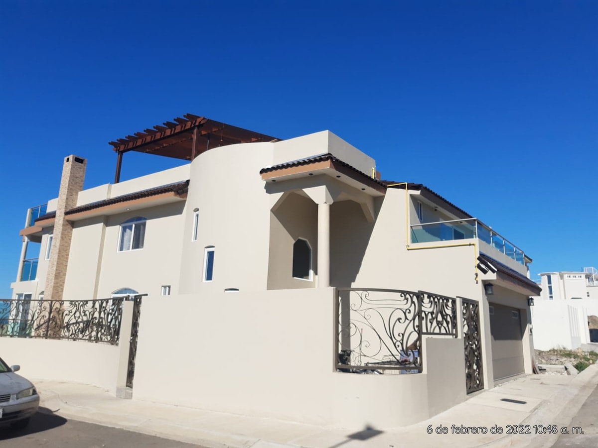 The exterior of a modern two-story house is showcased, featuring a smooth stucco facade and a terracotta-tiled roof. A spacious terrace with a wooden pergola is visible on the upper level, and decorative wrought iron fencing surrounds the property, complementing the clear blue sky.