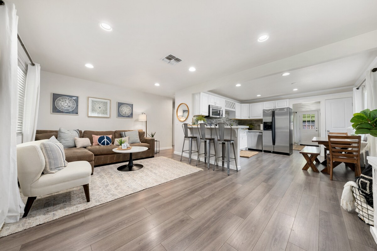 A spacious living area features a comfortable sectional sofa facing a coffee table, with decorative wall art enhancing the decor. A kitchen bar with seating is visible, along with modern appliances in the kitchen area. Natural light is welcomed through large windows adorned with light curtains.