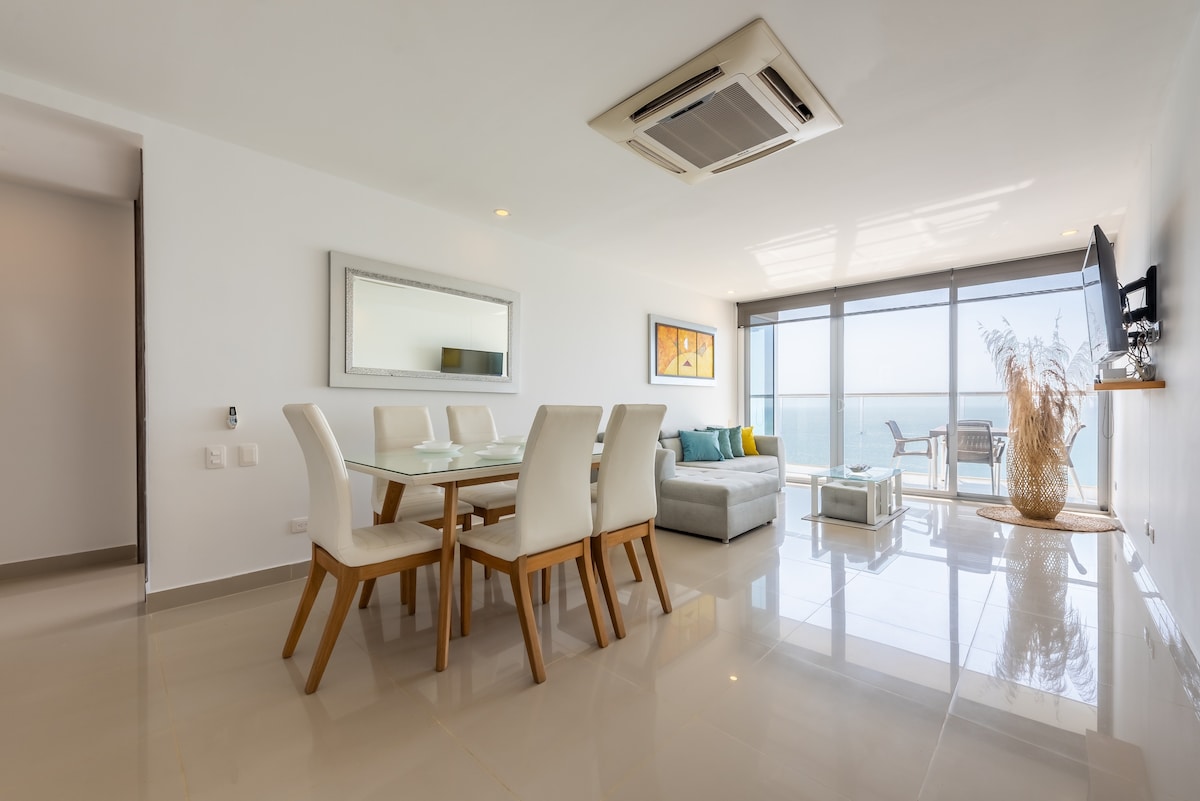 An airy living and dining area presents a glass dining table surrounded by white chairs. A comfortable sofa is situated nearby, with large windows offering views of the sea. A mounted TV and decorative accents contribute to a modern aesthetic.