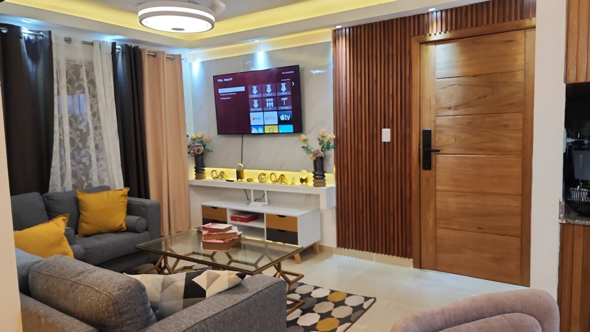 A stylish living area features a flat-screen TV mounted on a light-colored wall. A glass coffee table is centered on a patterned rug, surrounded by gray sofas adorned with yellow accent pillows. Soft curtains frame the windows, adding warmth to the space.