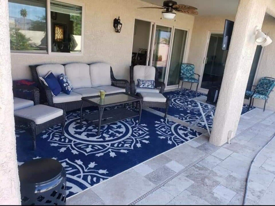 A covered patio area is displayed, featuring comfortable seating arranged around a central table. Decorative pillows add a touch of color to the light-toned furniture. A patterned rug complements the space, and large sliding doors provide access to the indoor area.