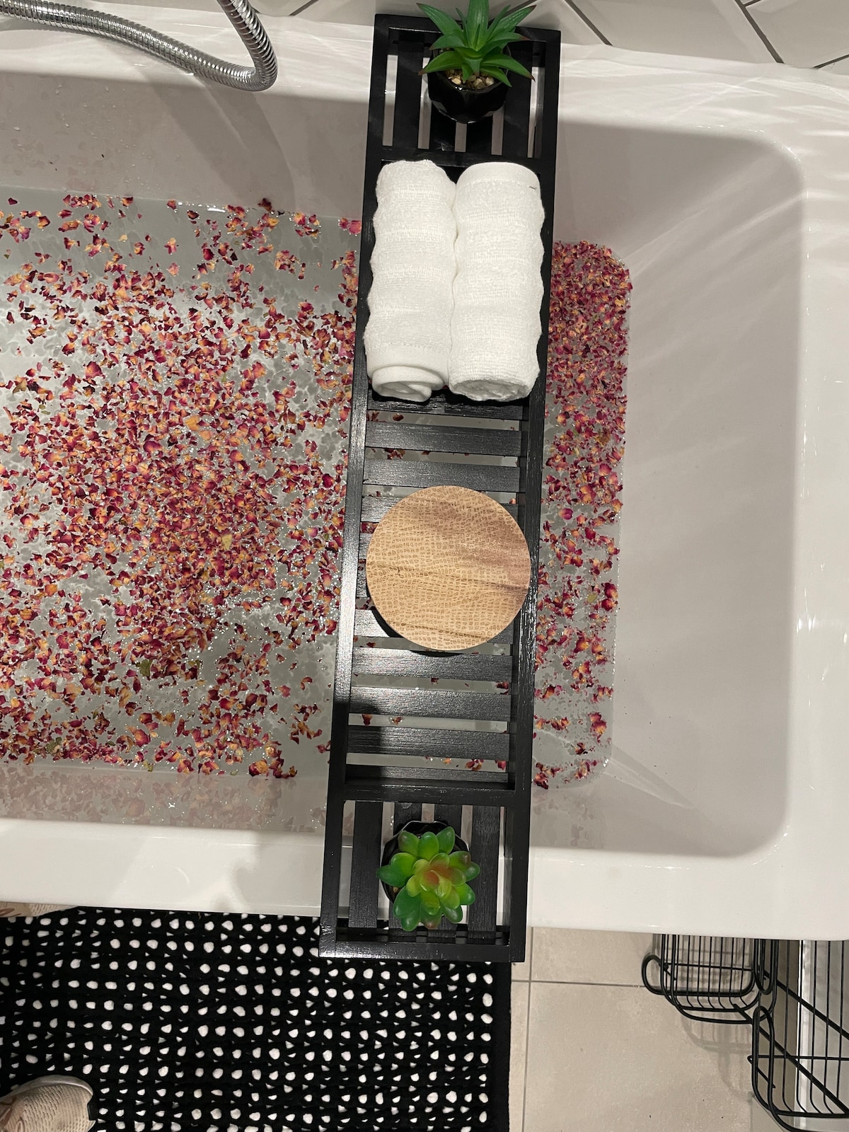 An elegant bath tray rests across the edge of a bathtub filled with scattered flower petals. Soft white towels are neatly stacked next to a small decorative plant and a circular wooden accent piece. The area is complemented by a black and white patterned rug.