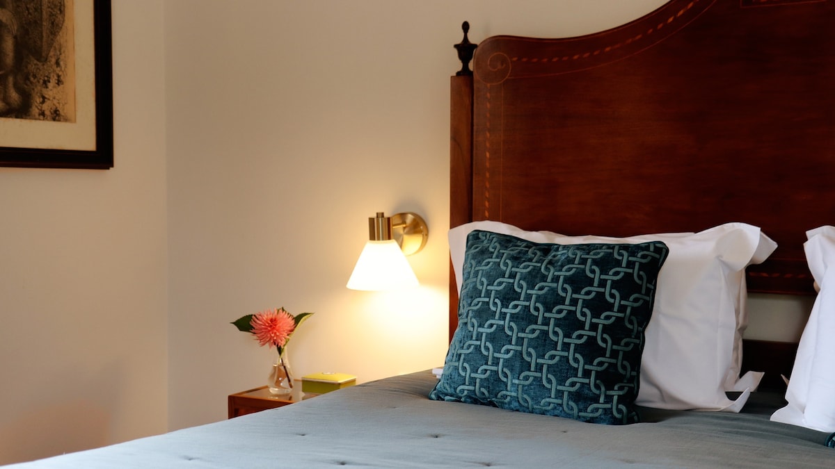 A cozy bedroom features a spacious bed with decorative pillows and a textured blanket. A bedside table holds a simple vase with a flower, while a modern wall sconce provides soft lighting.