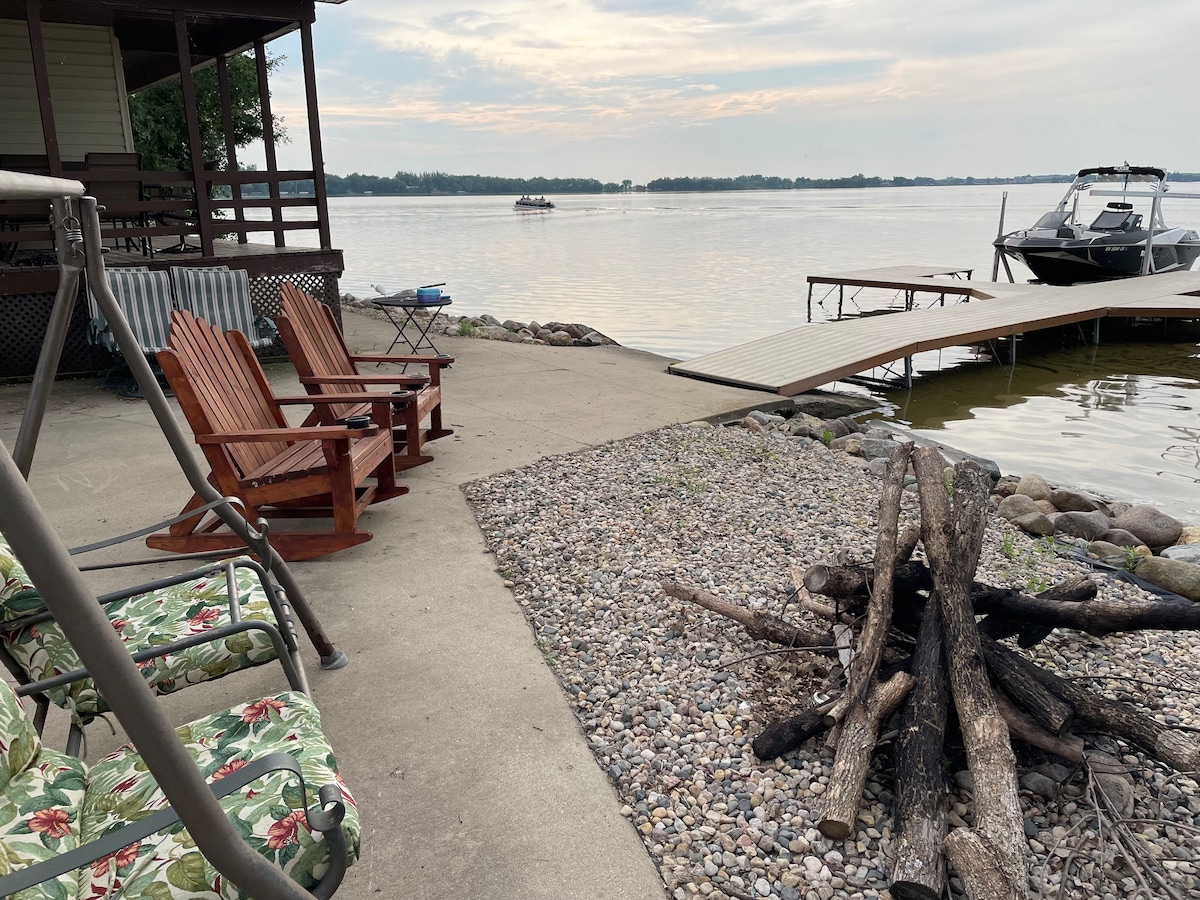 A serene lakeside area features a fire pit surrounded by rustic logs and a gravel patch. Two wooden rocking chairs are positioned nearby, with a swing chair visible in the foreground. A boat dock extends into the calm water, reflecting the gentle hues of the sky.