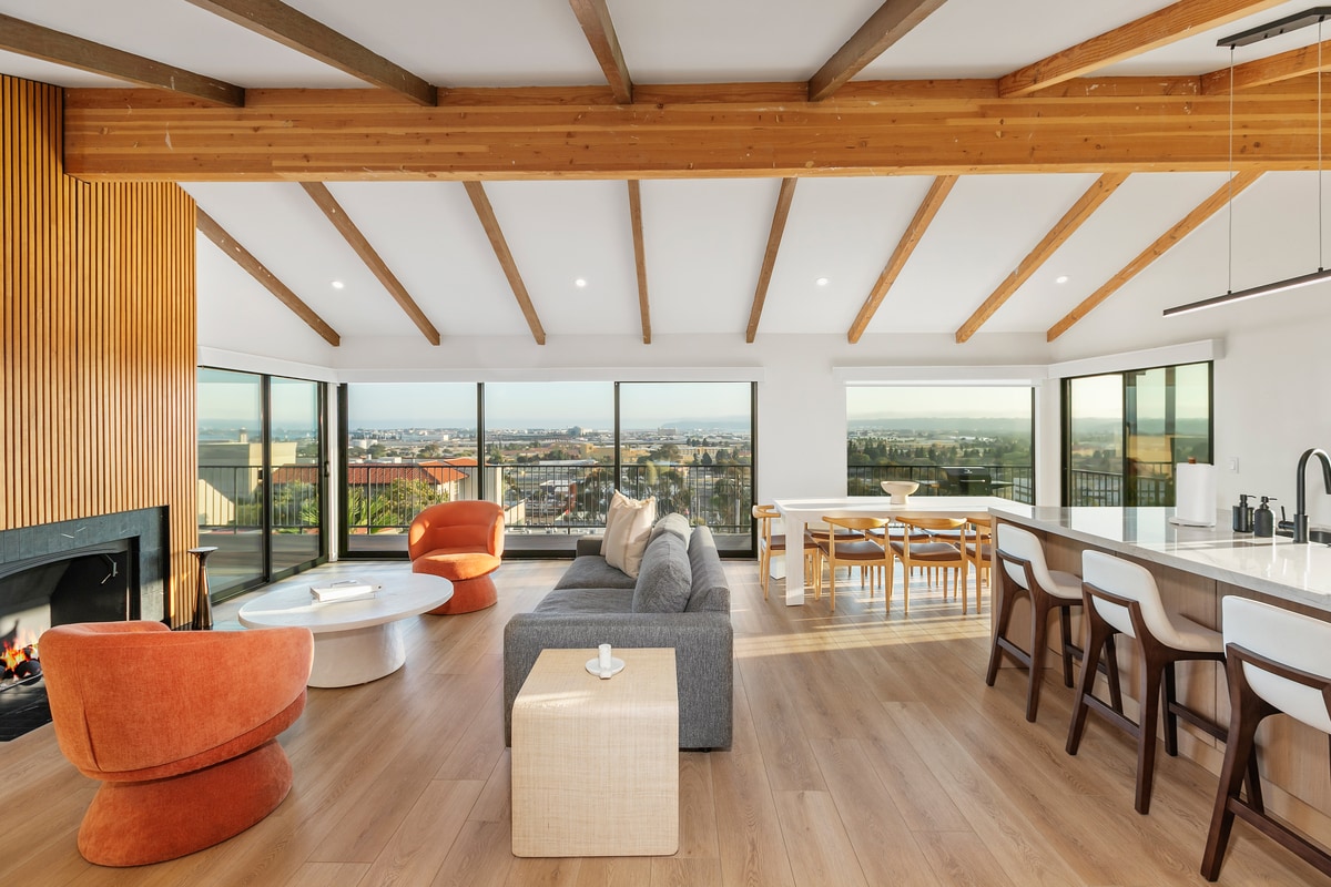 An open living area is showcased with vaulted wooden ceilings and large windows, allowing natural light to fill the space. A gray sofa and orange accent chairs are arranged around a white coffee table. A dining table is visible in the background, complemented by a modern kitchen.