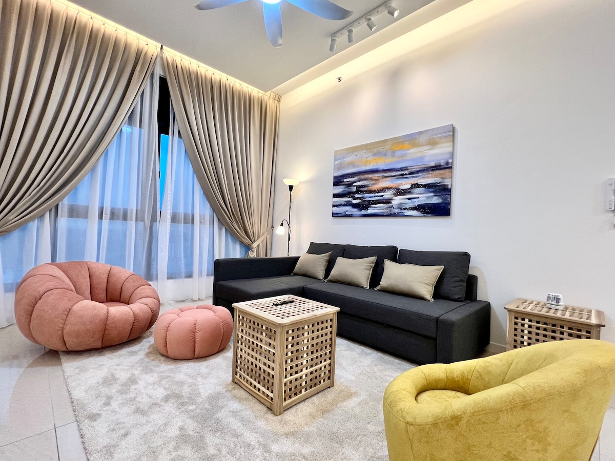 A welcoming living area is presented with a spacious, dark sofa and colorful accent chairs. Soft, round cushions are placed on a plush rug, while large windows dressed in flowing curtains allow natural light to fill the space, contributing to a relaxing atmosphere.