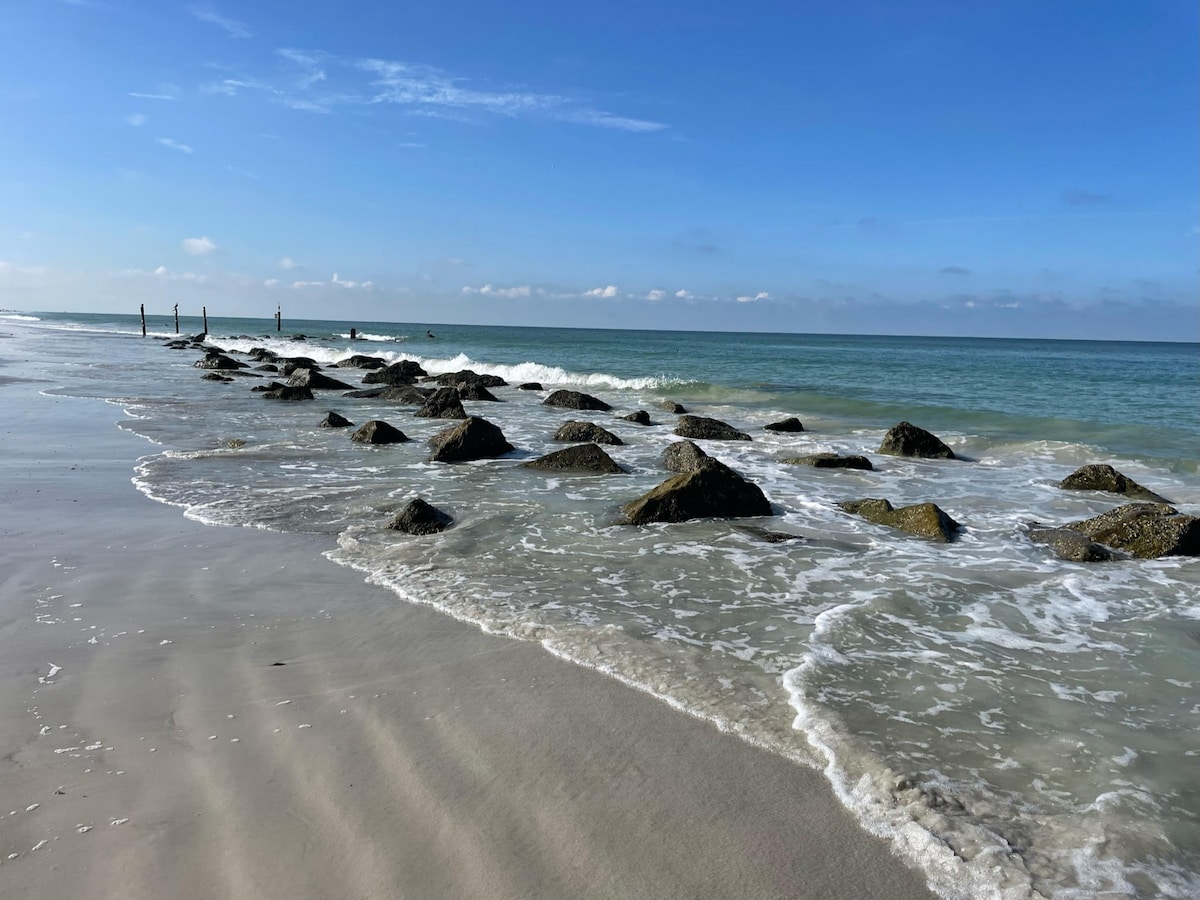 A serene beach scene is captured, featuring gentle waves lapping at smooth sand. Natural rock formations are visible just offshore, creating a textured contrast against the clear, blue waters and bright sky. The horizon is unobstructed, enhancing the calming ambiance of the coastline.