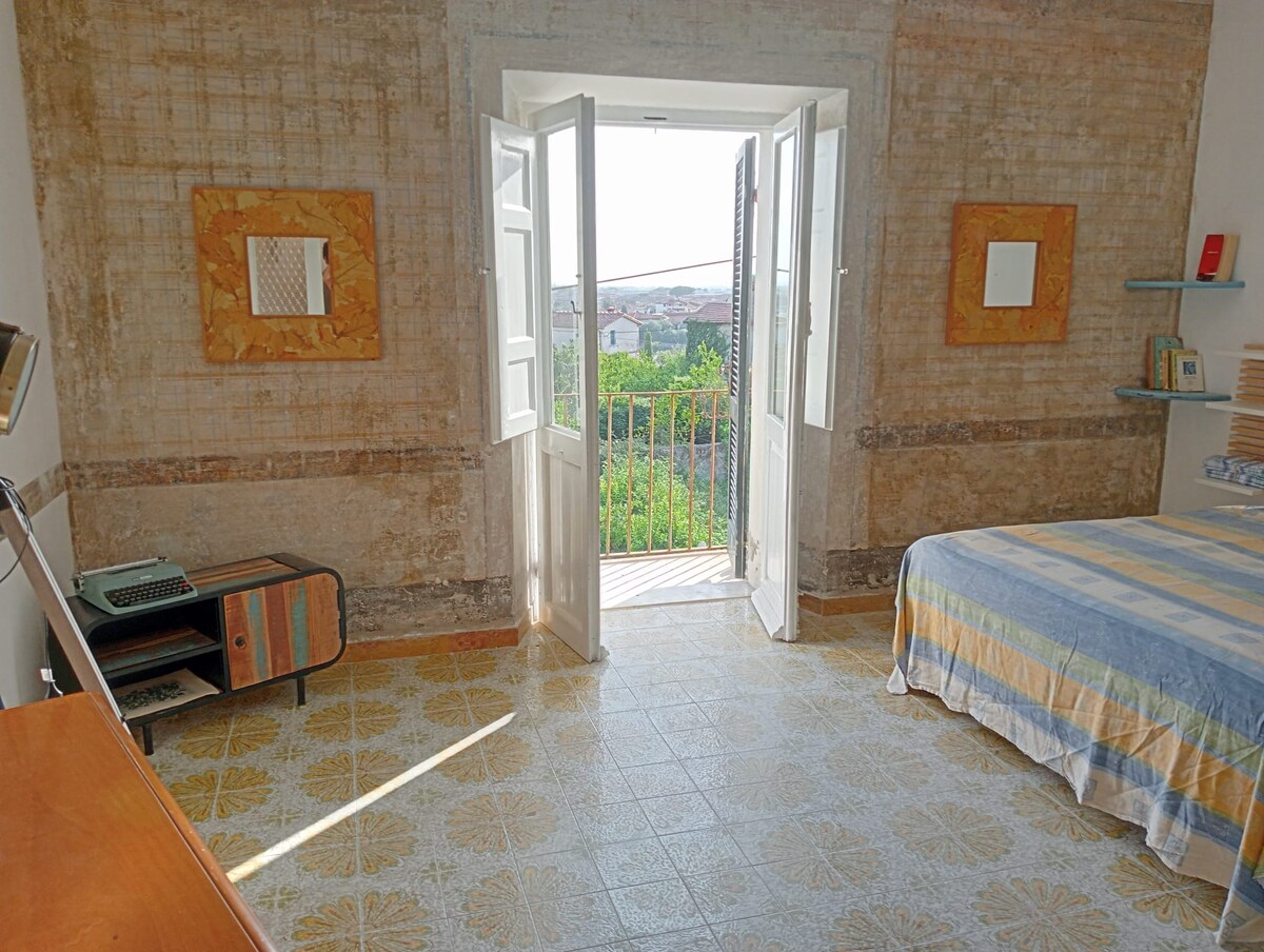 A bright bedroom features large double doors opening to a balcony, allowing natural light to fill the space. The room is adorned with textured walls, a colorful bedspread on a spacious bed, and vintage furniture, including a cabinet and a small writing desk.