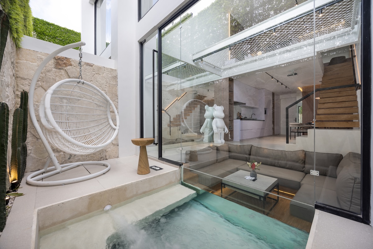 A stylish plunge pool is featured at the entrance, with clear water inviting relaxation. A hanging chair and a wooden side table are positioned beside the pool. Large glass doors provide a view into the open living space, showcasing modern furnishings and a light-filled ambiance.
