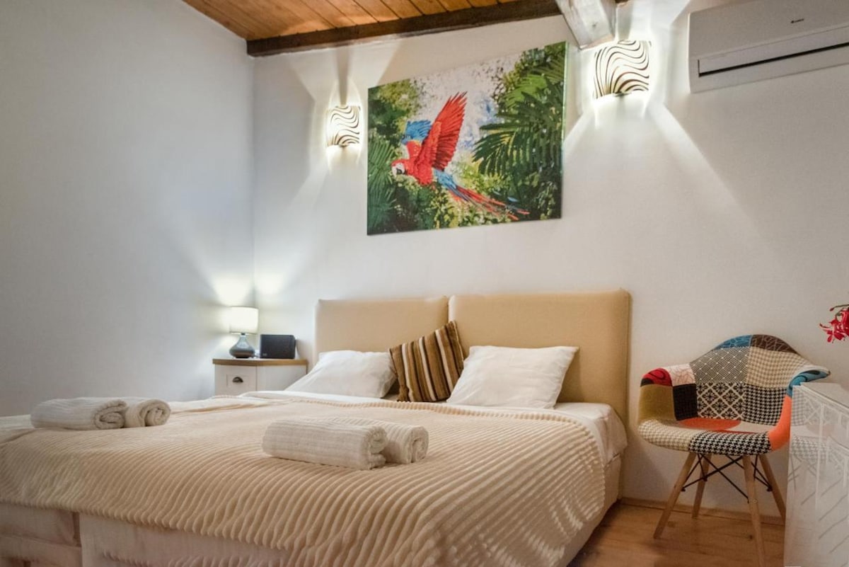 A spacious bedroom features a large bed adorned with neutral-colored linens. Decorative towels are neatly arranged. A vibrant artwork of a parrot hangs above the bed, complemented by soft lighting from wall sconces. A stylish chair adds a touch of character to the room.