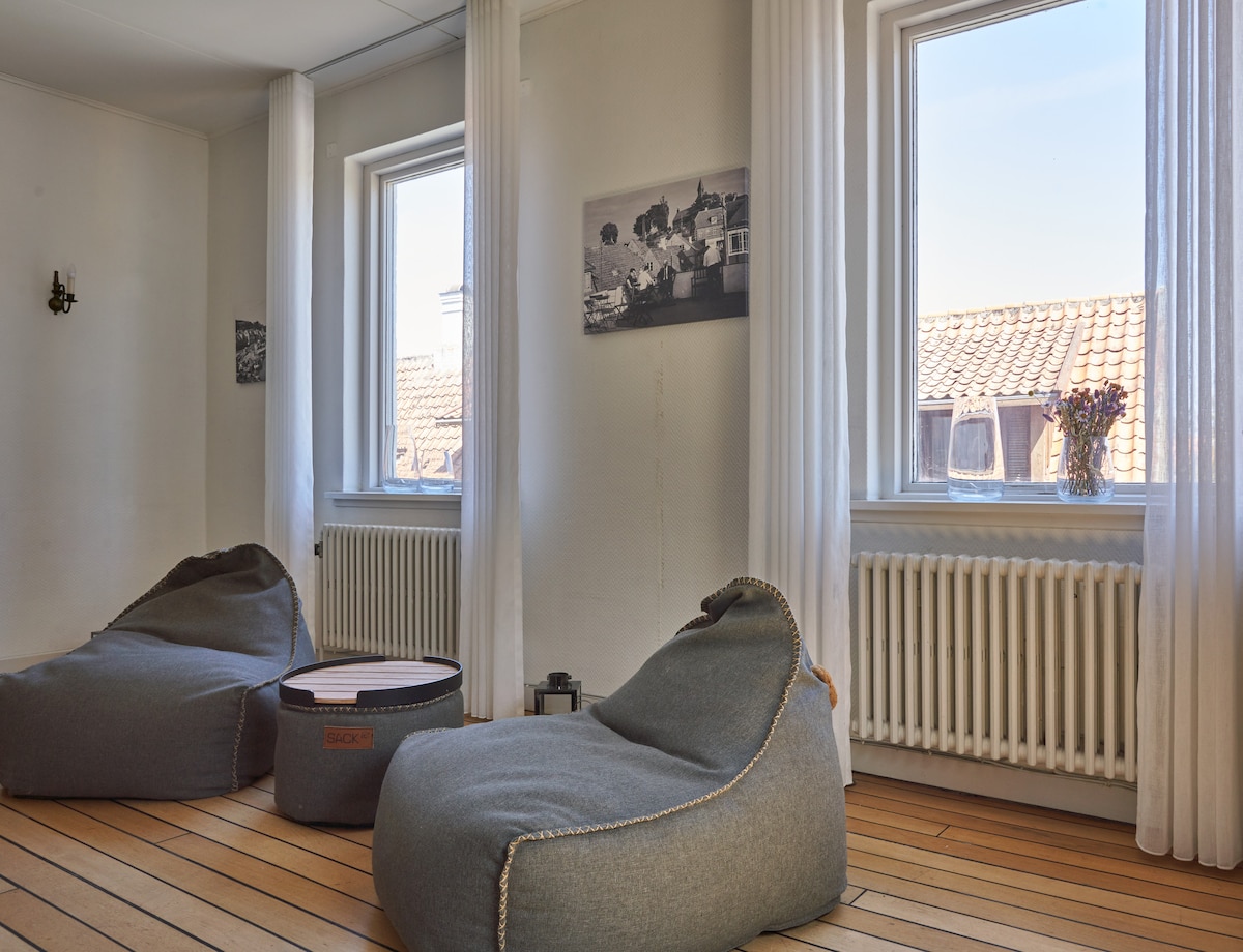 A bright and airy room features two comfortable bean bag chairs and a low circular table. Natural light streams through two large windows adorned with sheer white curtains. A black-and-white photograph hangs on the wall, complementing the room's warm wooden flooring.