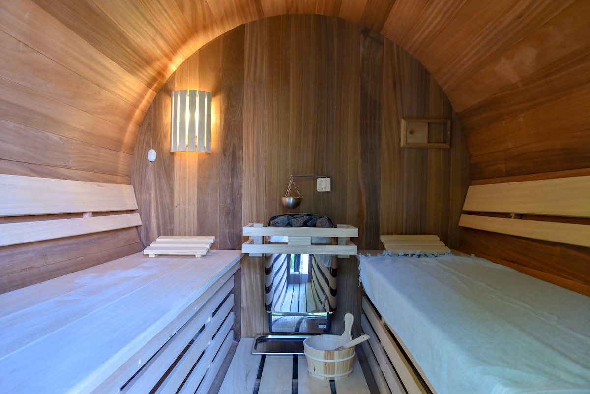 The interior of a wooden sauna features two spacious lounging benches, designed for relaxation. Soft ambient lighting is provided by a wall fixture, while a small bowl is ready for use. The warm wood grain creates a calming atmosphere throughout the room.