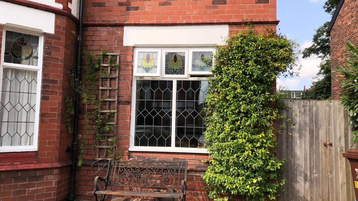 A charming exterior view showcases a brick wall adorned with decorative stained glass windows. Lush greenery surrounds the window, and a wooden bench is positioned nearby. A wooden trellis adds texture to the scene, enhancing the inviting and natural ambiance of the entrance.