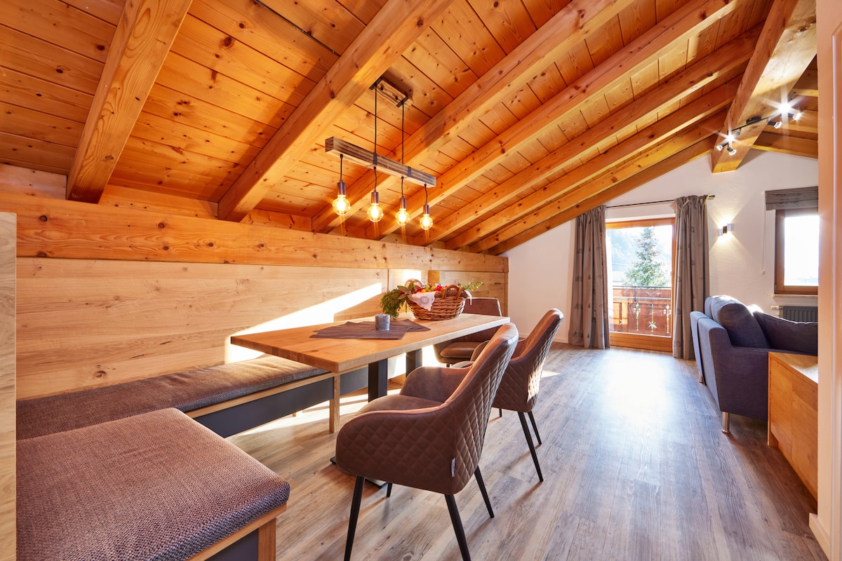 A spacious living and dining area showcases a wooden ceiling with exposed beams. A large table is surrounded by dark upholstered chairs, complemented by a cozy seating area with a sofa. Natural light streams through a window, offering outdoor views and enhancing the warm wood tones.