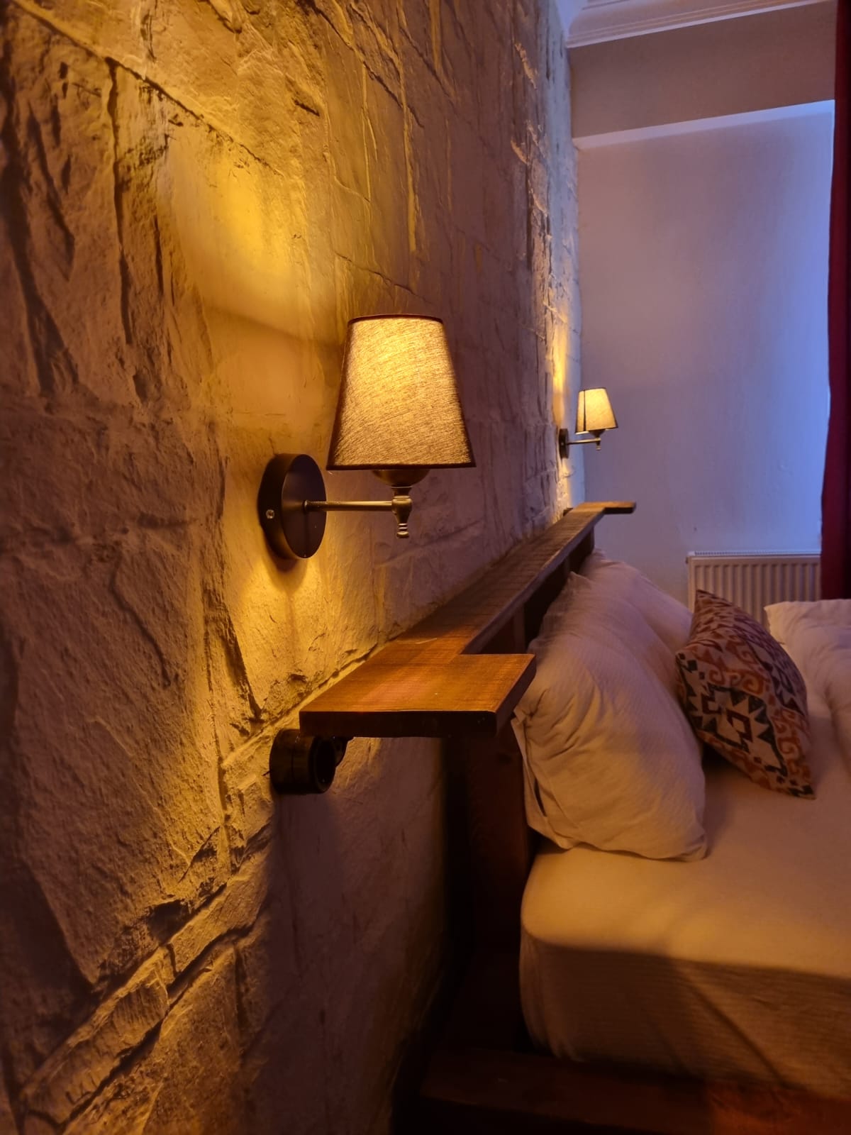 A spacious bed is positioned against a textured stone wall, illuminated by soft, warm lights from matching sconces. Decorative cushions add a touch of style, while natural light filters through a nearby window dressed in deep red curtains.