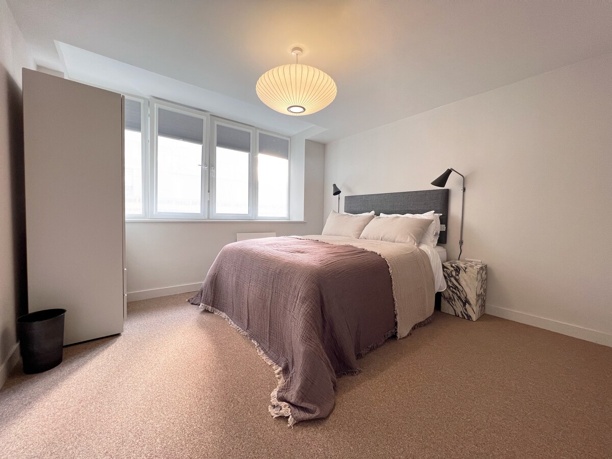 A spacious bedroom is equipped with a large bed featuring a soft, muted blanket. A modern light fixture hangs from the ceiling, illuminating the room. Large windows allow natural light to fill the space, while minimalist furniture, including a cabinet and a bedside table, completes the setting.