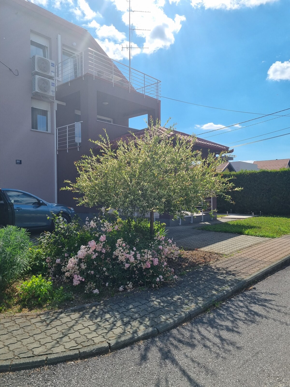 An exterior view showcases a multi-story building with air conditioning units mounted on the side. Lush greenery and a flowering bush enhance the entrance, while a paved pathway leads up to the property. Clear skies and sunlight add to the inviting atmosphere.