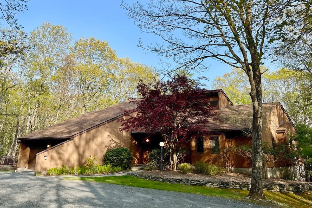 A spacious home is set within a natural landscape, featuring earthy-toned exterior walls and a sloped roof. Lush trees frame the entrance, complemented by a vibrant red-leafed shrub. A gravel driveway leads up to the house, enhancing the serene woodland setting.