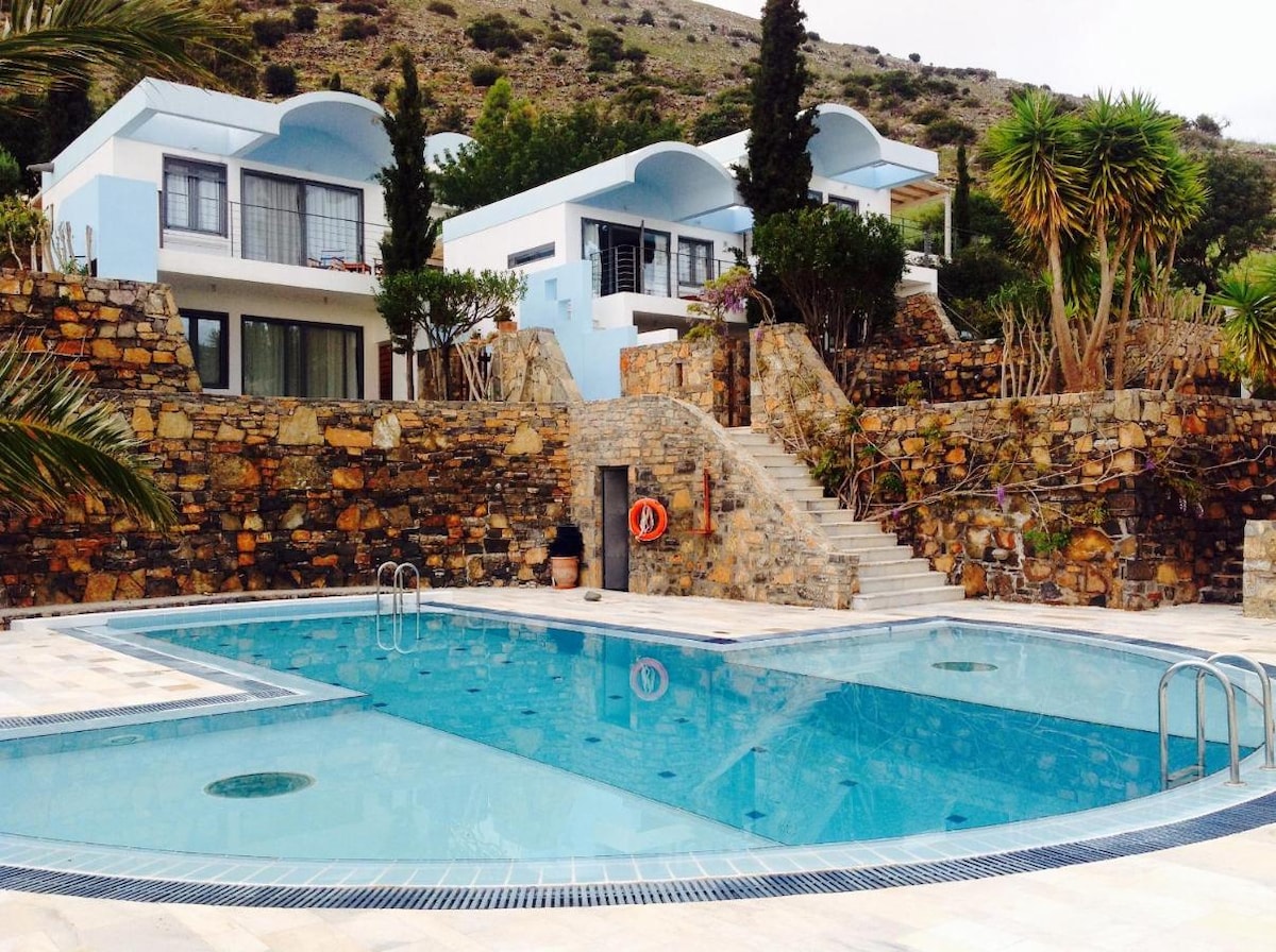 A central swimming pool is surrounded by natural stone features and well-maintained greenery. The pool reflects the sky, creating a calm atmosphere. Villas are positioned on elevated terrain, with steps leading to accessible areas, enhancing the inviting setting.