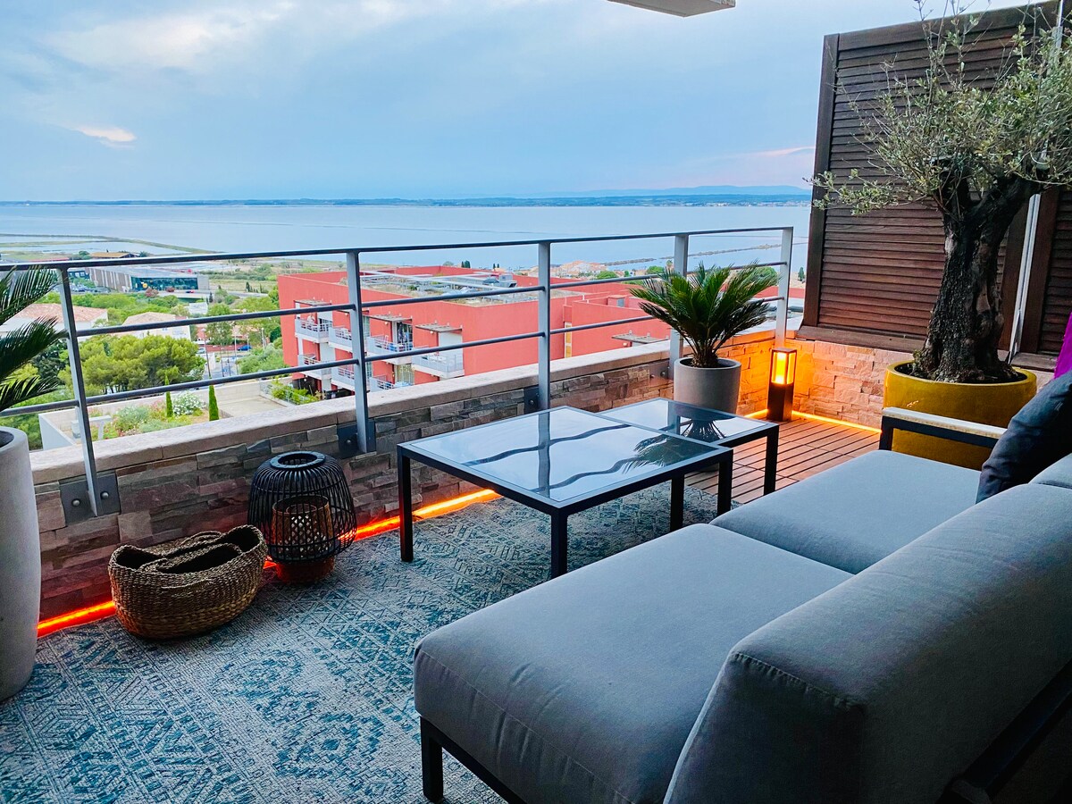 An outdoor terrace features comfortable seating and a glass table, with a backdrop of expansive views over the Thau lagoon and Mediterranean Sea. Decorative plants complement the space, while subtle lighting along the floor adds ambiance during twilight hours.