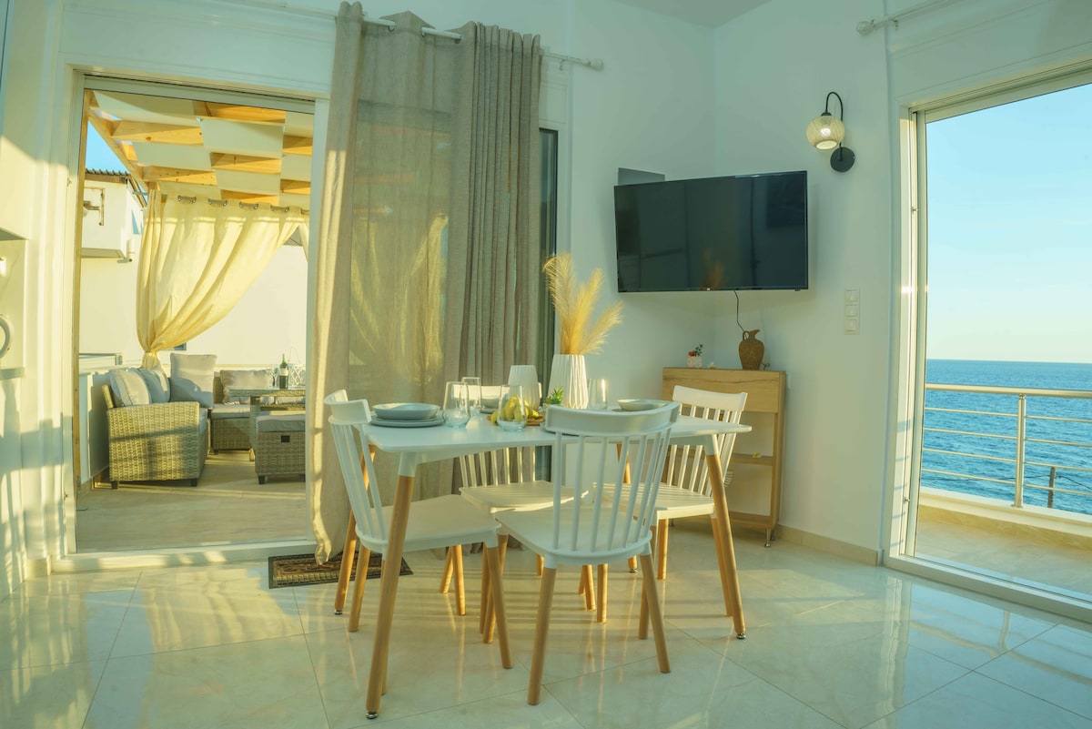 A light-filled dining area features a round table set for two, surrounded by white chairs. A modern television is mounted on the wall. To one side, expansive glass doors lead to a balcony, providing views of the ocean and outdoors.