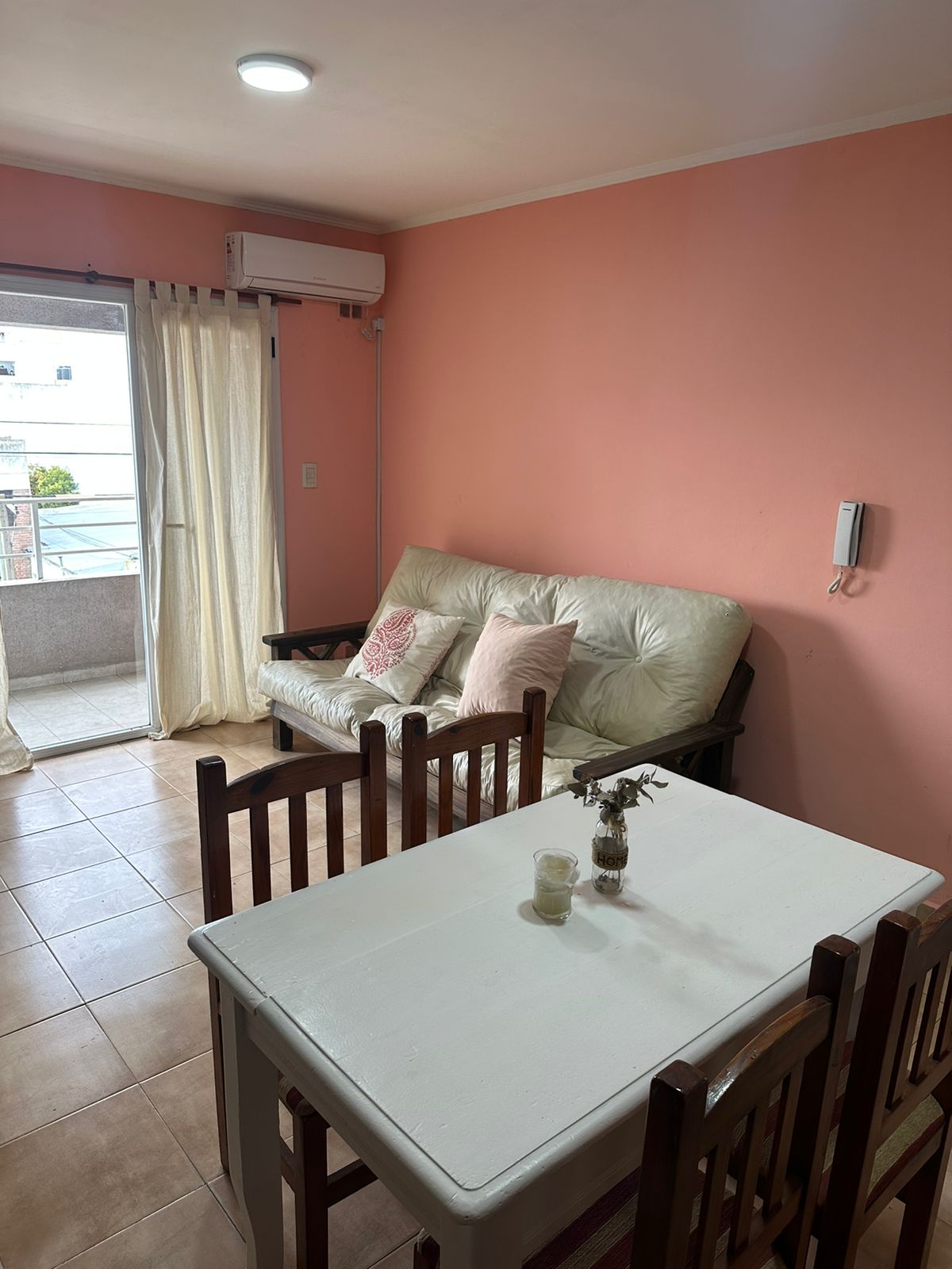 A light-filled living space features a comfortable futon adorned with decorative pillows, positioned near a window that leads to the balcony. A small dining table with wooden chairs sits opposite, creating an inviting area for meals or gatherings. Soft peach walls add warmth to the room.