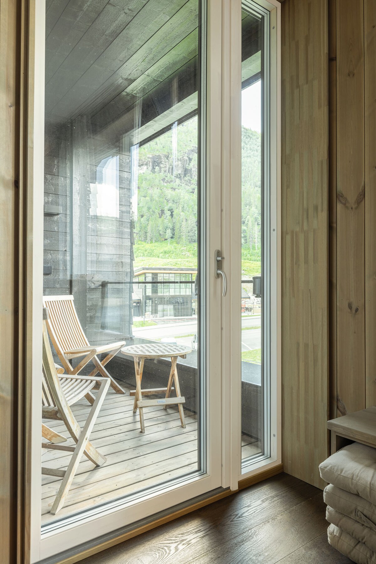 A balcony area is visible through a glass door, showcasing a pair of wooden chairs and a small folding table. Natural light filters in, illuminating the warm wood paneling and providing a view of the surrounding hillside.