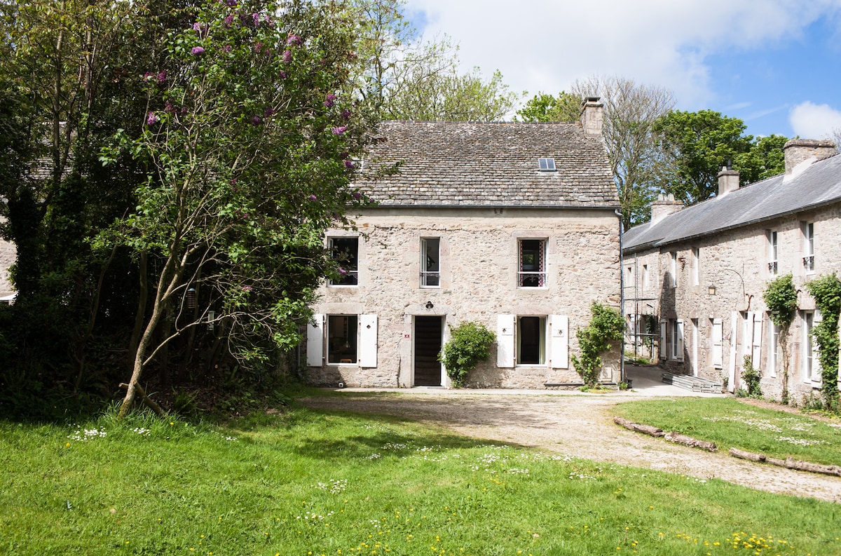 The exterior of a stone house is shown, featuring a green lawn and a pathway. Sunlight illuminates the facade, highlighting the windows adorned with shutters. Lush greenery surrounds the property, enhancing the natural setting. Two adjacent buildings are visible, creating a comfortable courtyard.