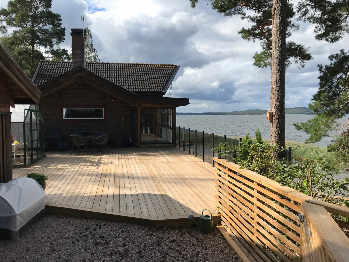 A spacious deck made of wood extends towards the water, providing a clear view of the surrounding nature. Comfortable seating arrangements can be seen, with soft shadows created by nearby trees. The exterior features a log cabin with large windows reflecting the tranquil lake and cloudy sky.
